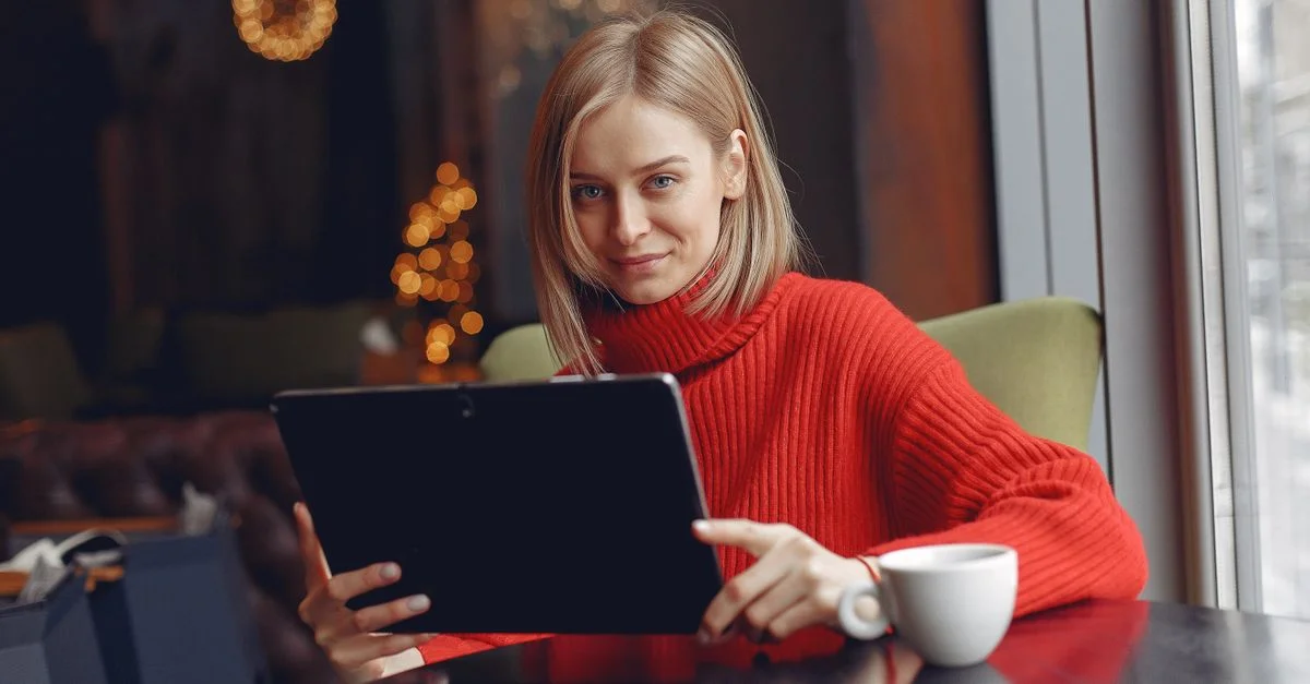 Femme dans un café convivial avec une tablette et un café, portant un pull rouge.